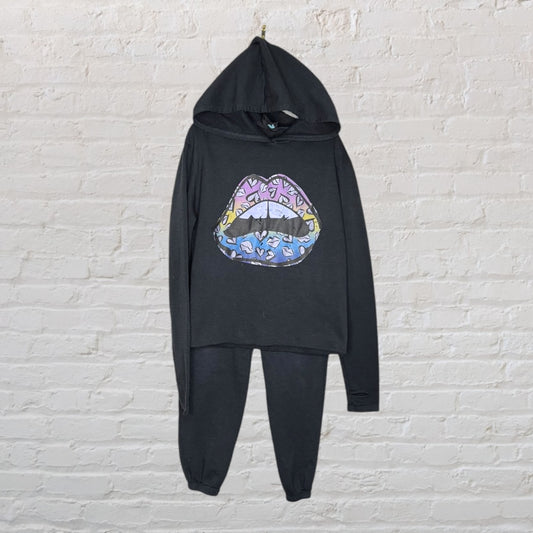 Black hoodie and pant set with a multicolored graphic of lips on the top, hanging against a brick wall.