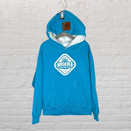 Blue hoodie from Muskoka Bear Wear with front pocket and logo print, displayed on a hanger against a brick wall.