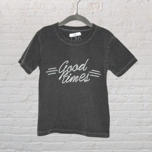 Charcoal T-shirt with cursive 'Good Times' text printed in white on a hanger against a white brick wall.