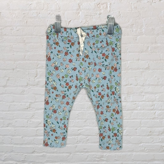 Light blue baby leggings featuring a multicolored floral pattern, hanging on clips against a white brick background.