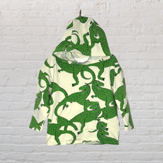 Kids' cream hoodie with green alligator print, featuring long sleeves and a hood, hanging against a white brick wall.