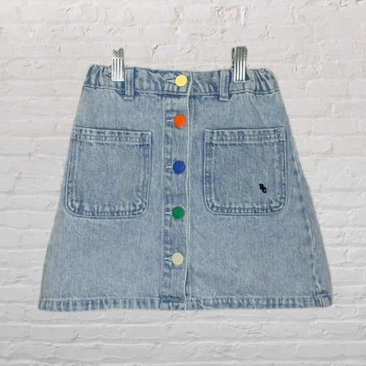 Light blue denim skirt with multicolored buttons and front pockets, displayed against a white brick background.