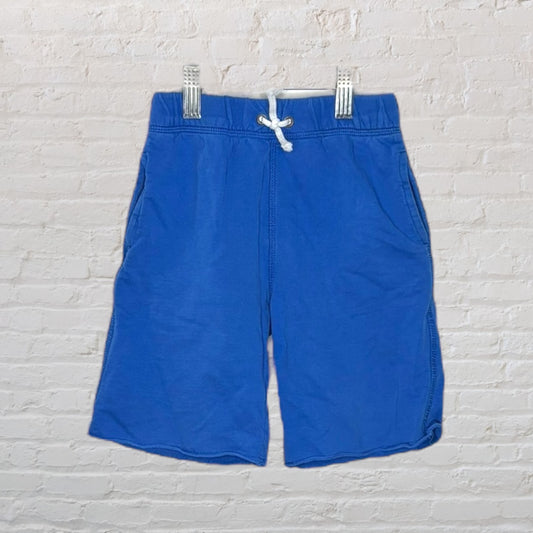 Blue casual shorts with a white drawstring and side pockets, hanging against a brick wall.