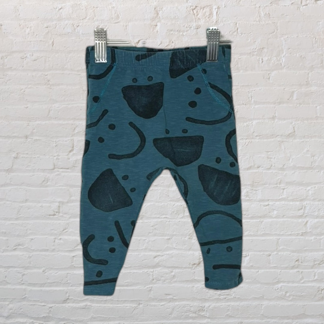 Teal baby leggings with black abstract patterns, hanging on clips against a white brick wall.