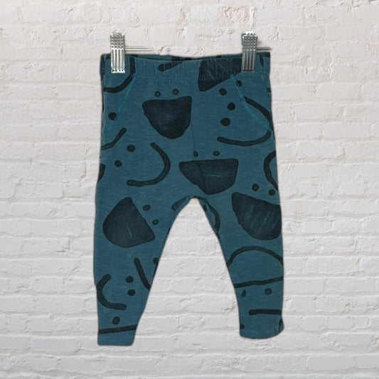 Teal baby leggings with black abstract patterns, hanging on clips against a white brick wall.