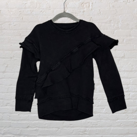 Black long-sleeve sweater with diagonal ruffle detail, displayed on a hanger against a white brick wall.