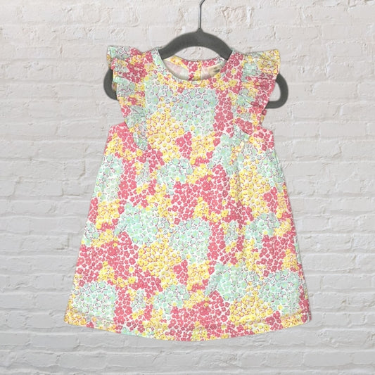 A sleeveless dress with floral patterns in pastel colors and ruffle sleeves, displayed on a hanger against a white brick wall.