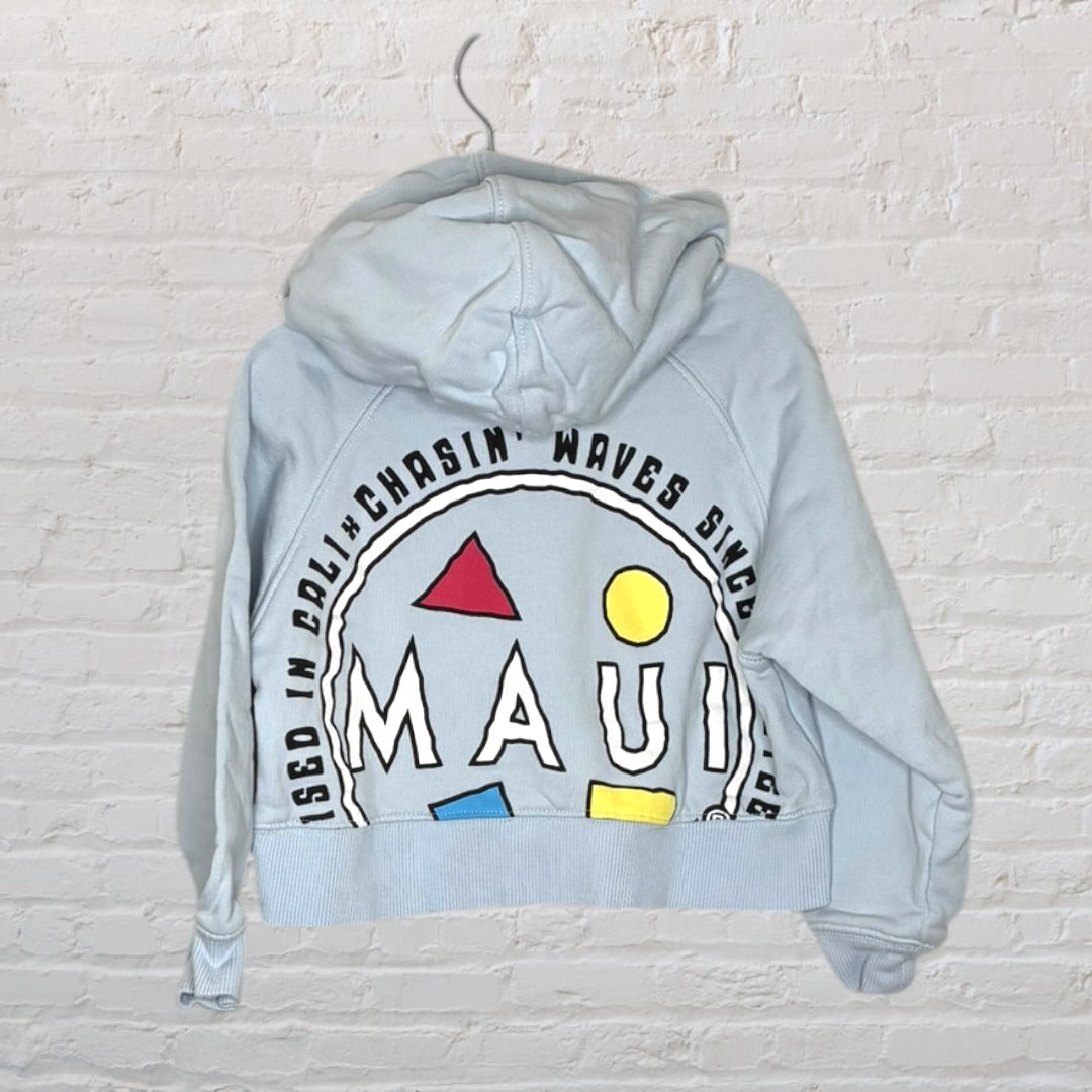 Light blue hoodie with a back graphic featuring 'Maui' and colorful geometric shapes, hanging against a white brick wall.