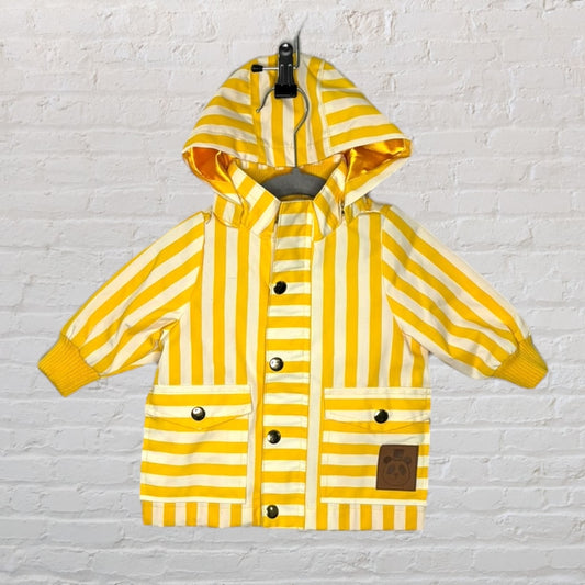 Yellow and white striped hooded raincoat for kids, featuring buttons and front pockets, hanging against a brick wall.
