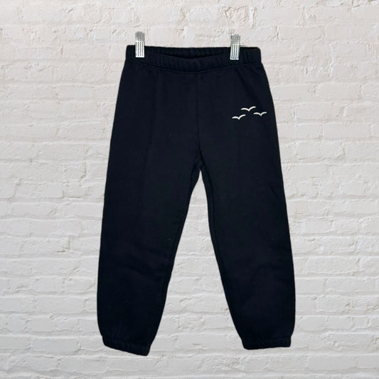 Black joggers with elastic waistband and bird embroidery on the upper leg, hanging against a brick wall.