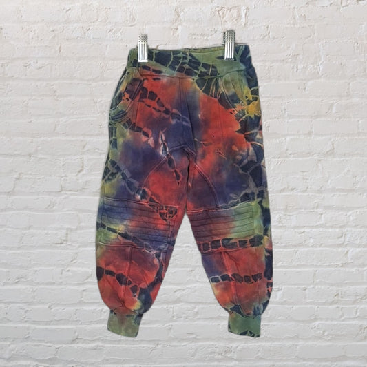 Multicolour tie-dye jogger pants with elastic cuffs and pocket details, displayed against a white brick wall.