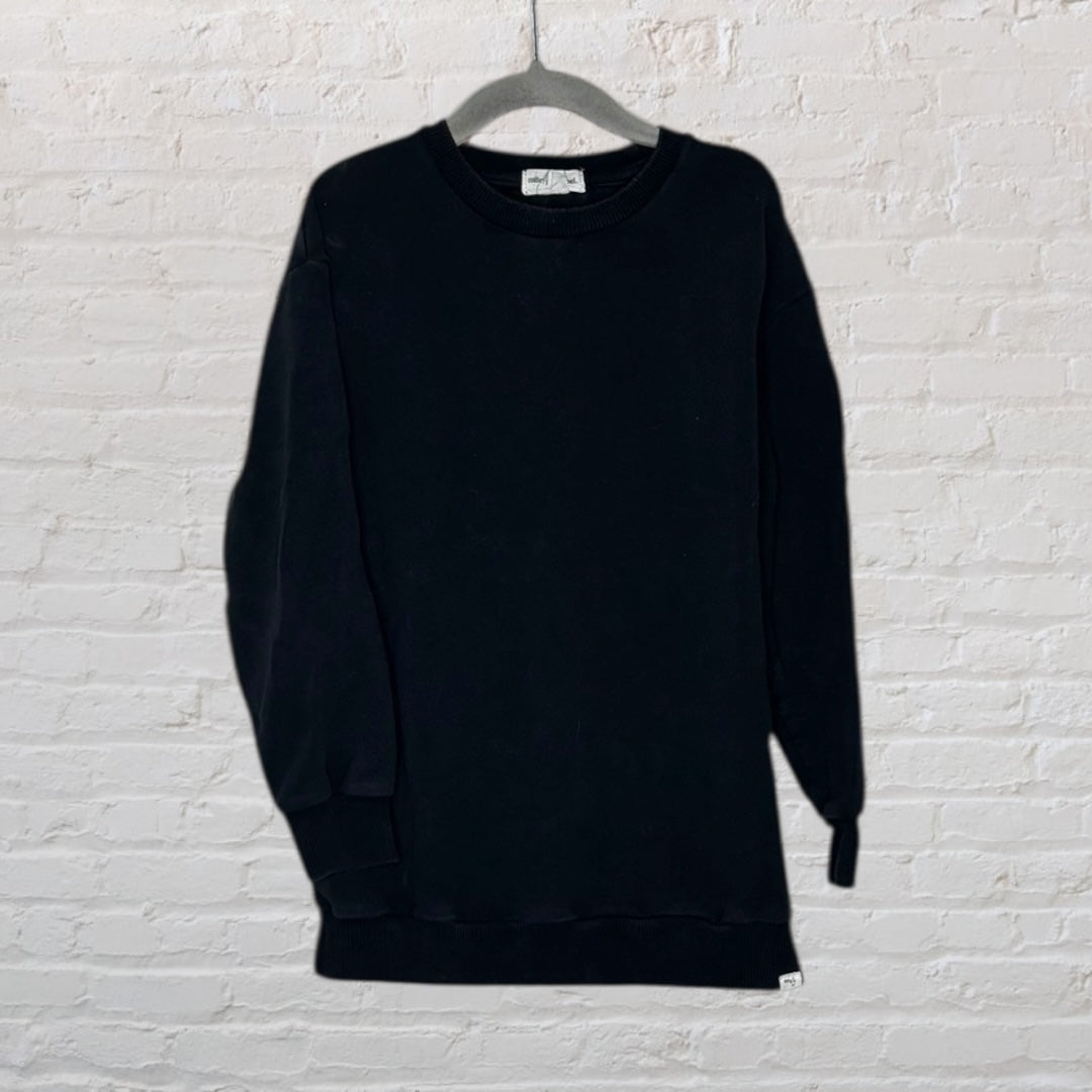 A plain black sweatshirt with a classic fit, displayed on a hanger against a white brick wall.