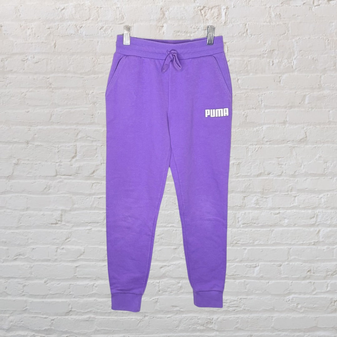 Purple Puma sweatpants with a drawstring waist and logo on the left thigh, hanging against a white brick wall.