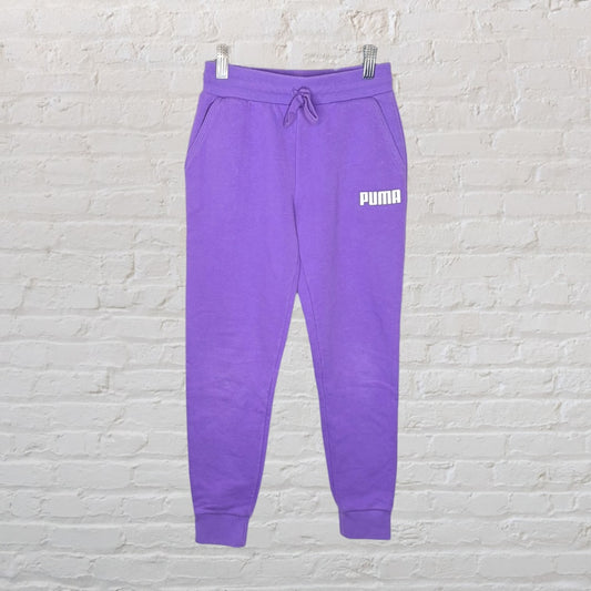 Purple Puma sweatpants with a drawstring waist and logo on the left thigh, hanging against a white brick wall.