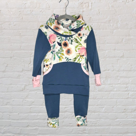 Children's blue and floral hoodie with matching pants featuring pink accents, displayed on a hanger against a white brick wall.