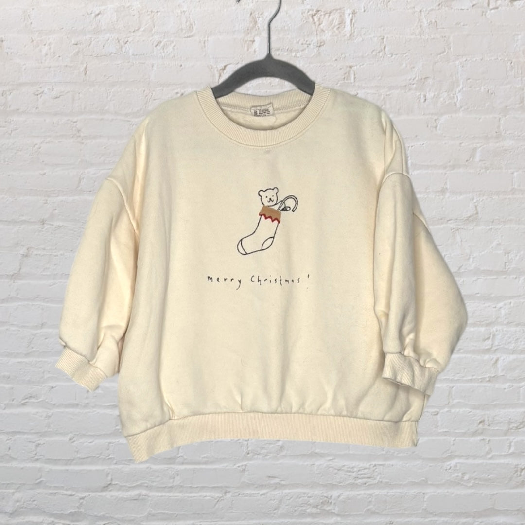 Cream sweatshirt with a cute bear in a stocking and 'Merry Christmas!' text, hanging on a hanger.