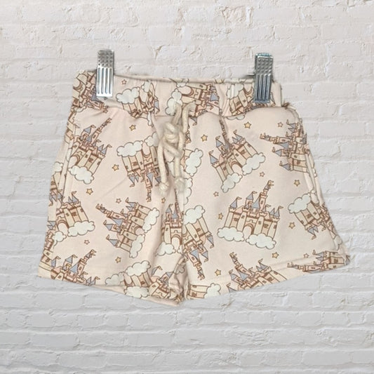 Beige shorts with a colorful castle and clouds print, featuring a drawstring waist, clipped on a white brick background.