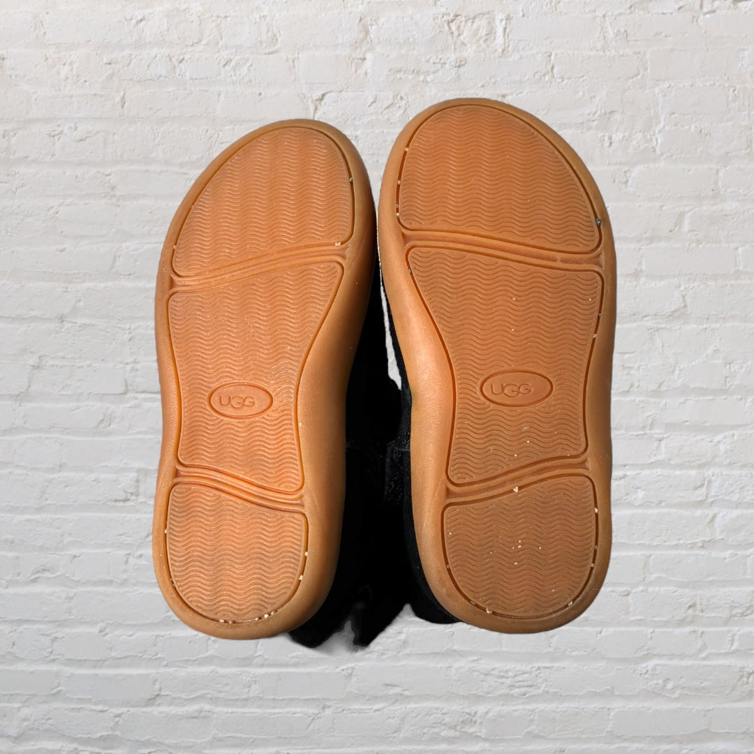 Black UGG boots with shearling lining and brown sole, side view against a brick wall background.