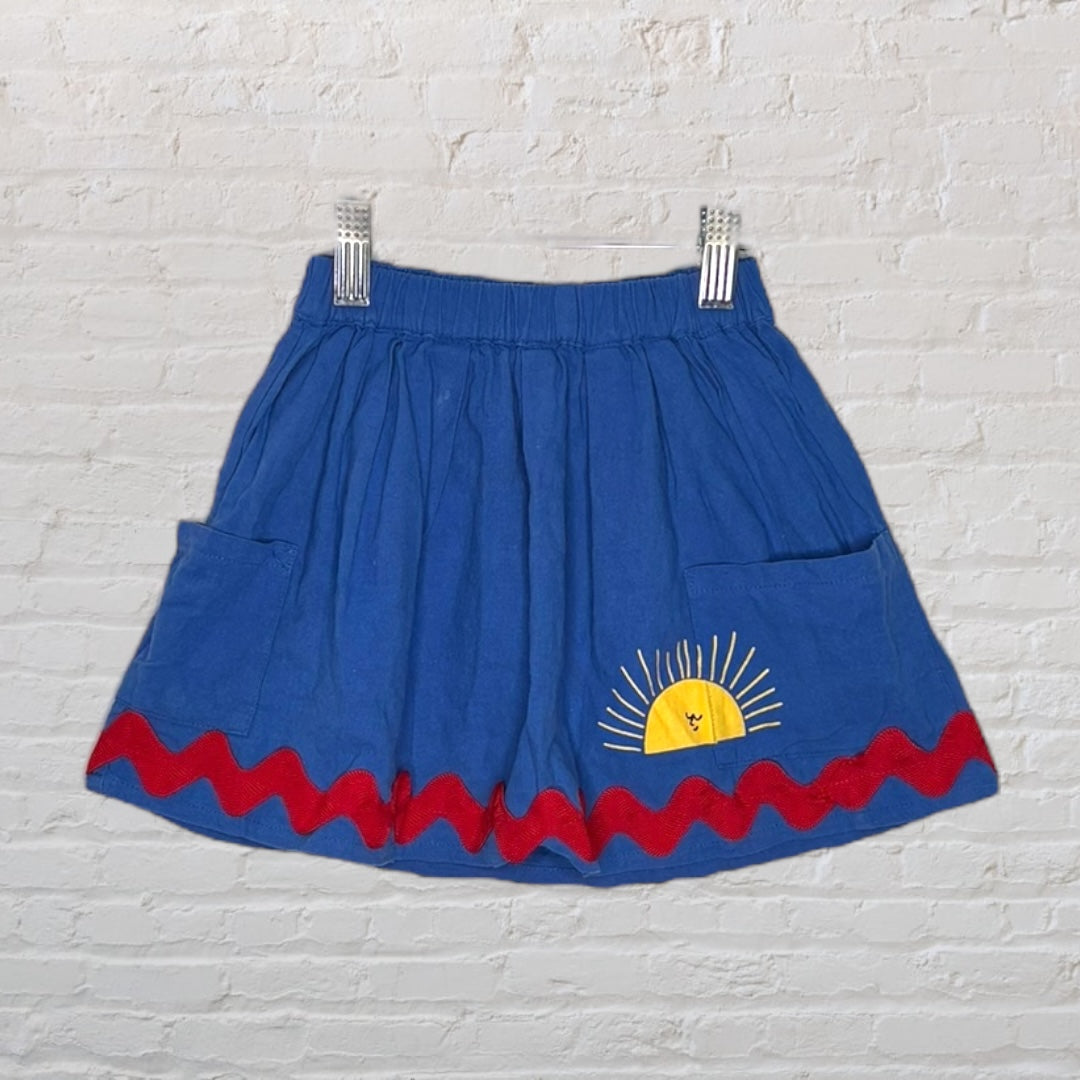 Blue skirt with sun embroidery and red wavy trim, featuring side pockets, displayed on a hanger against a white brick wall.