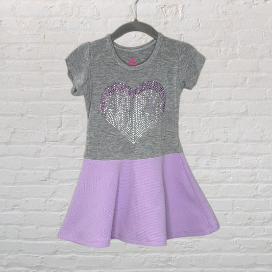 Two-tone dress with grey top featuring a sequined heart and lavender skirt portion, hanging on a hanger against a white brick wall.