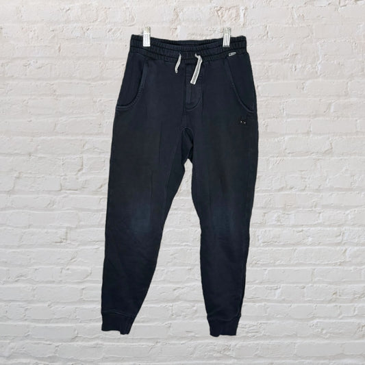 Black casual drawstring sweatpants with pockets and elastic cuffs, hanging against a white brick wall.