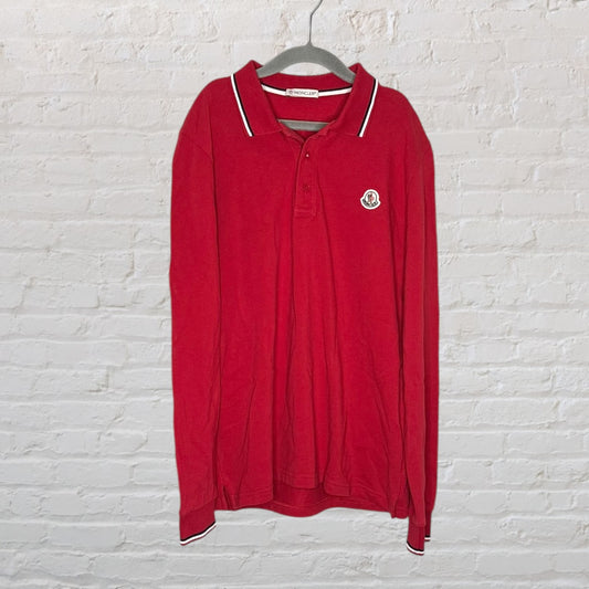 Red Moncler long sleeve polo shirt with white-tipped collar, hanging on a hanger against a brick wall.