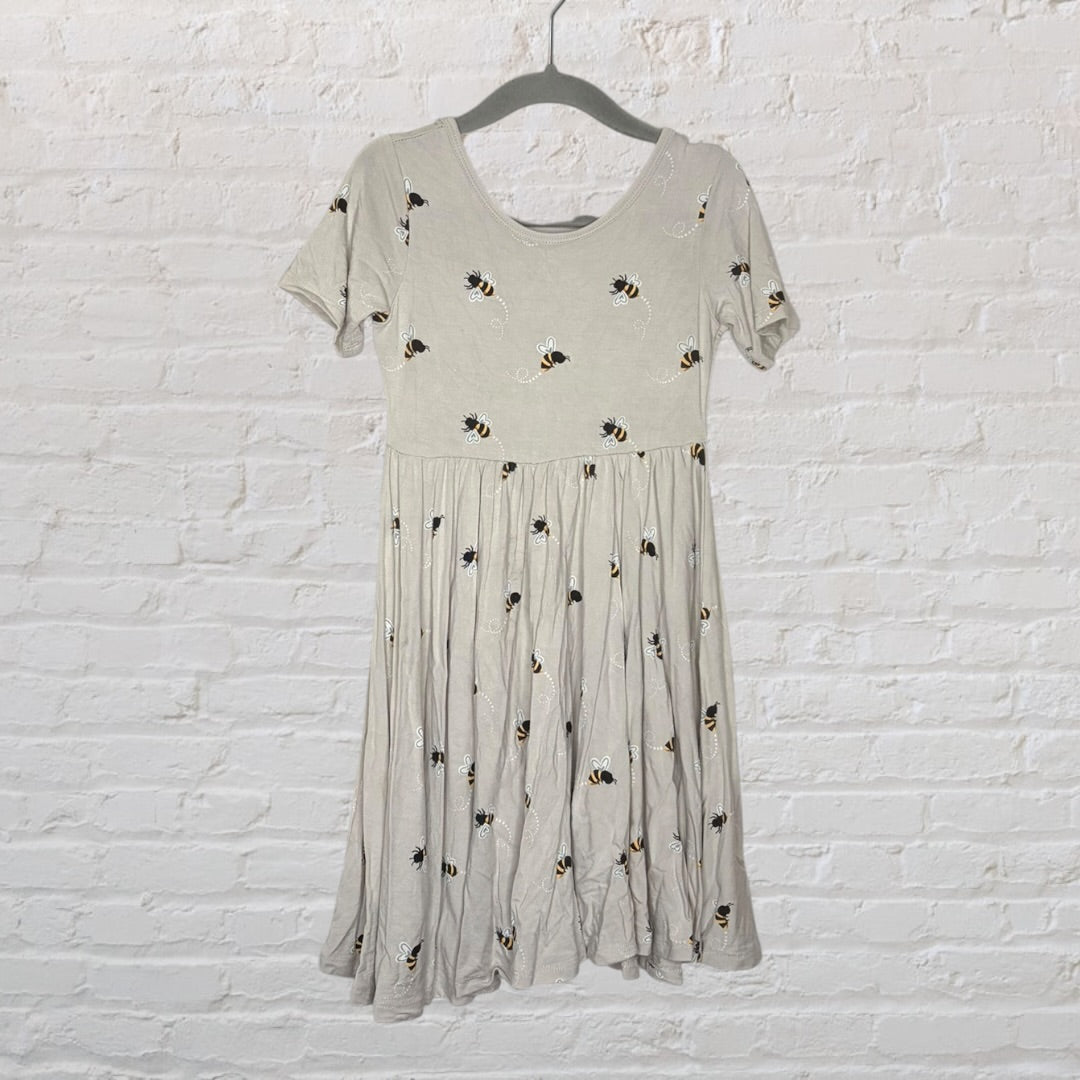 Light grey skater dress with bee patterns, hanging on a hanger against a white brick wall.