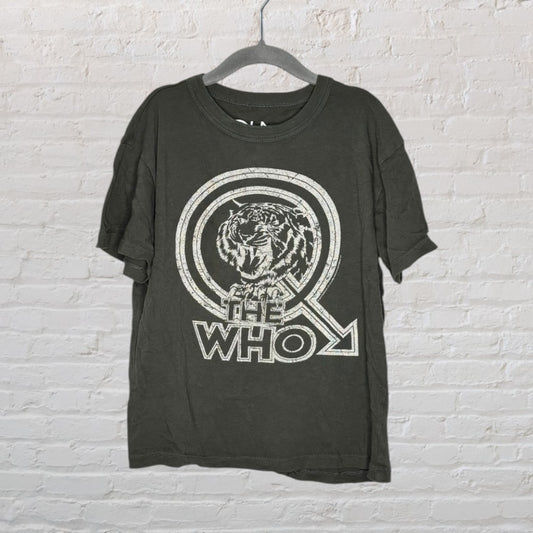 Black T-shirt with 'The Who' graphic featuring a tiger and arrow design, hanging on a gray hanger against a white brick wall.