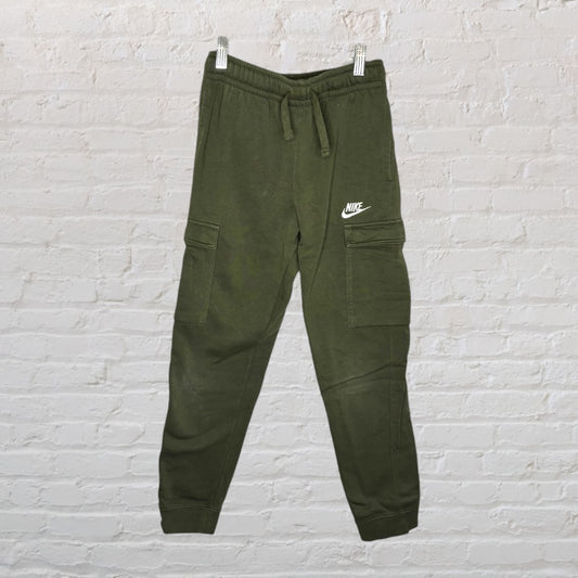 Olive green Nike cargo jogger pants with side pockets and drawstring waist, displayed hanging against a brick wall.