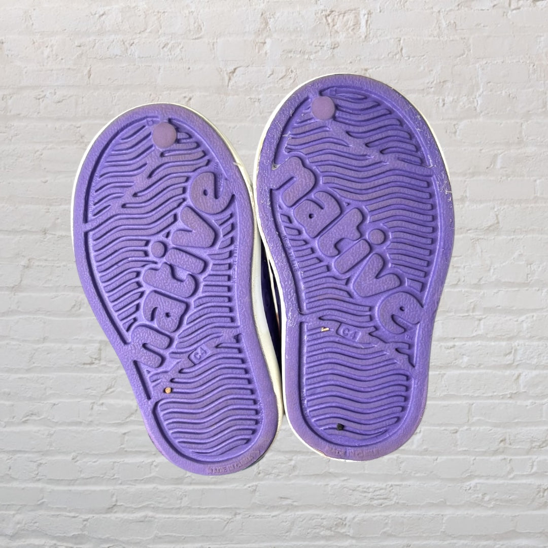 Purple perforated slip-on shoes with white sole, designed by Native, against a white brick background.