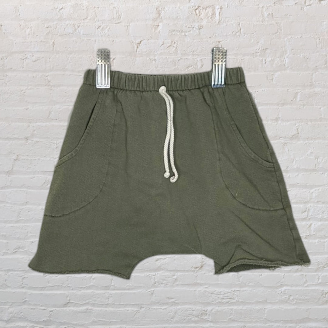 Olive green casual shorts with drawstring waist and pockets, clipped on a hanger against a brick wall.