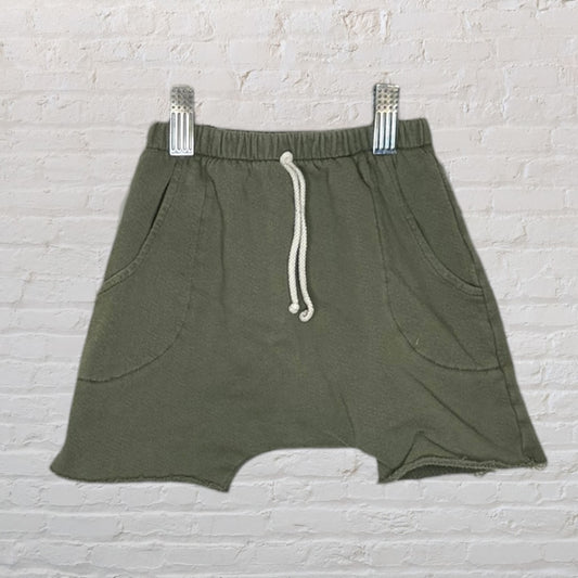 Olive green casual shorts with drawstring waist and pockets, clipped on a hanger against a brick wall.