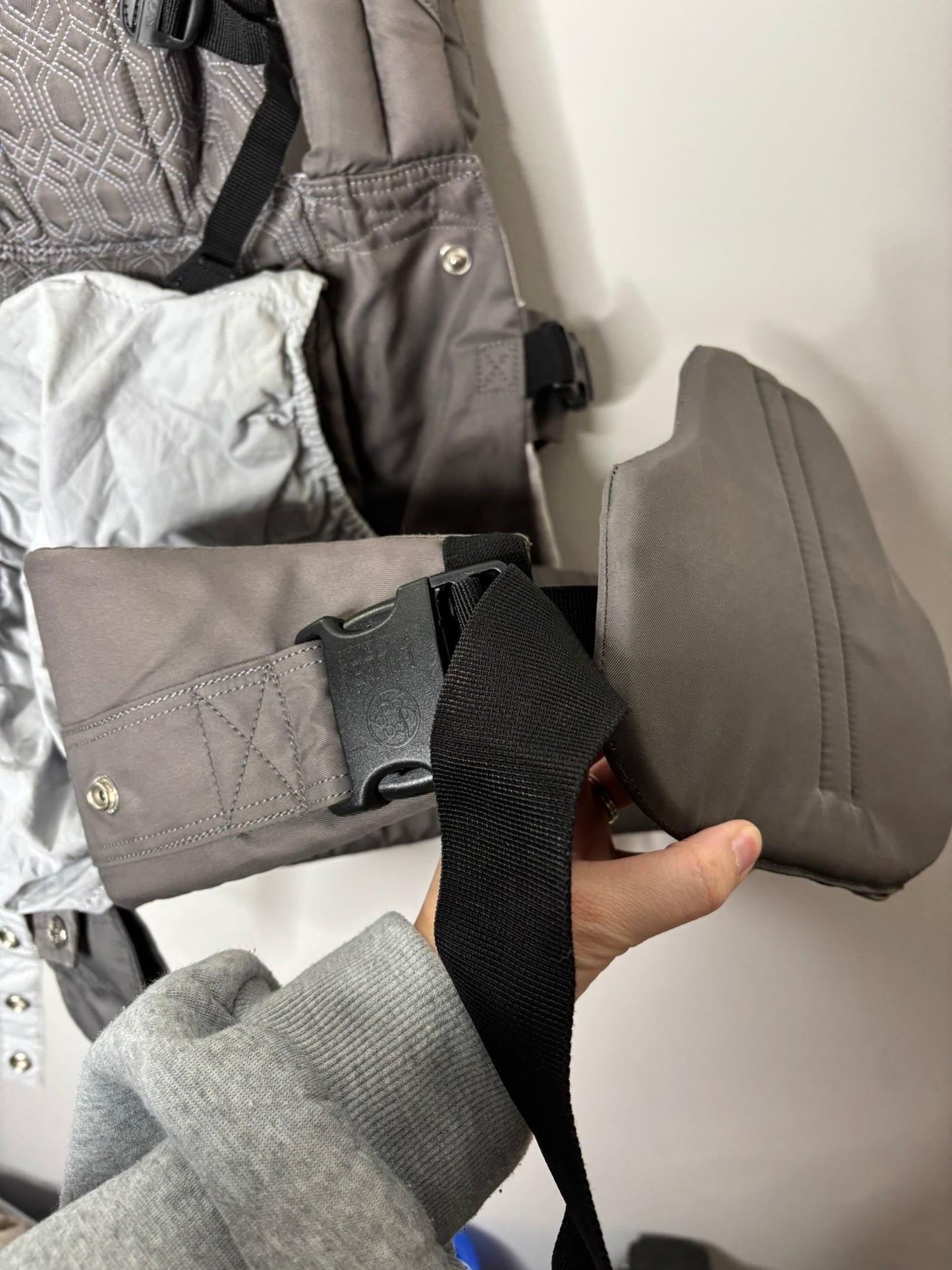 Gray ergonomic baby carrier with multiple straps and support, hanging on a hanger.