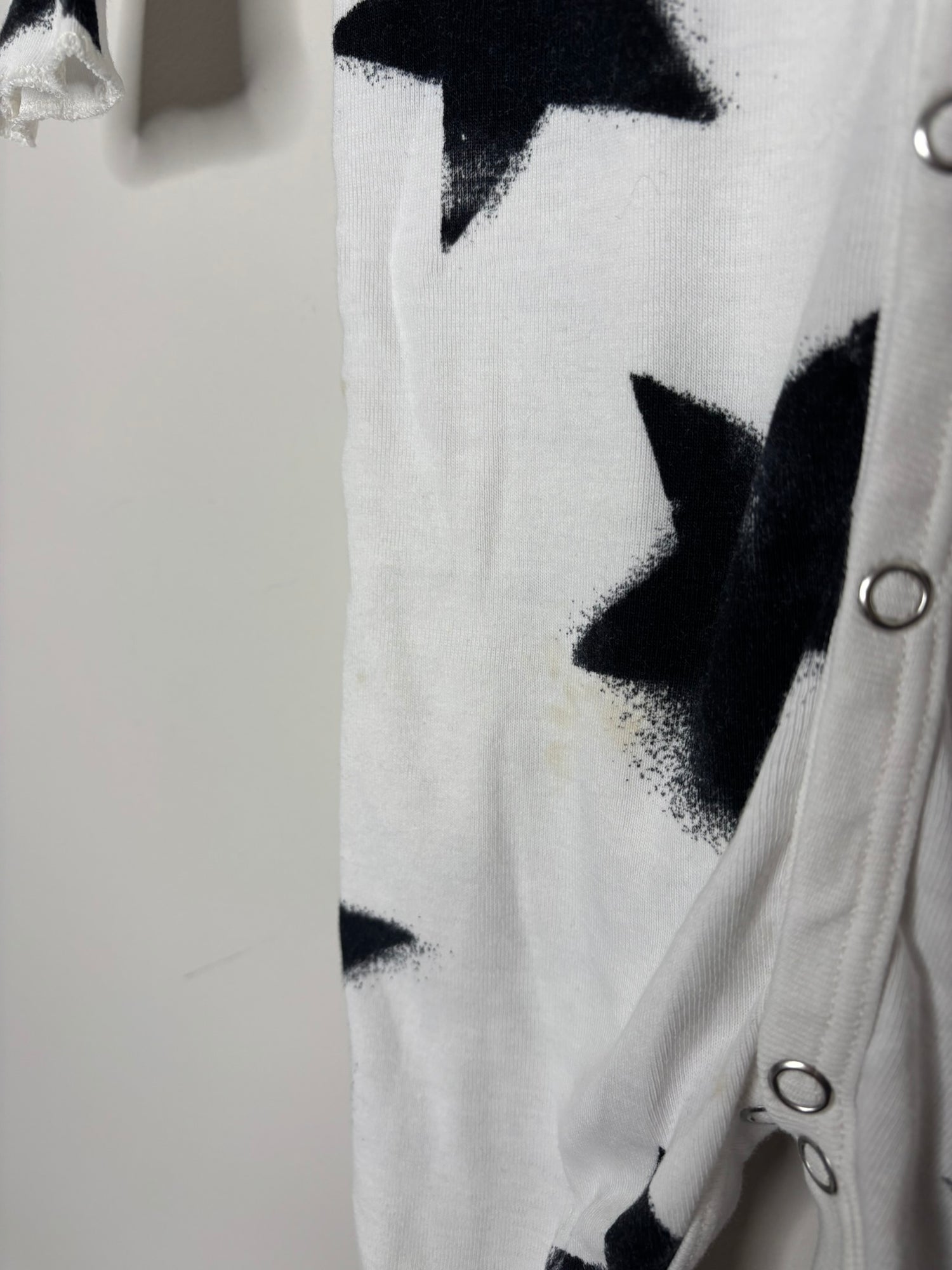 White baby onesie with black star print, featuring snap buttons down the front, hanging on a hanger against a brick wall.