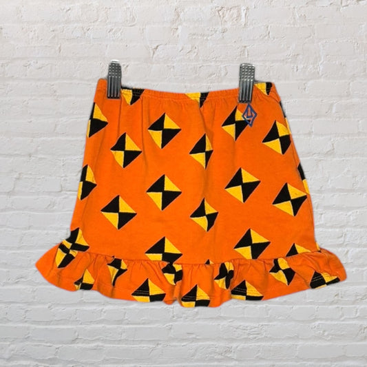 Orange skirt with black and yellow geometric pattern and ruffle hem hanging against a white brick wall.