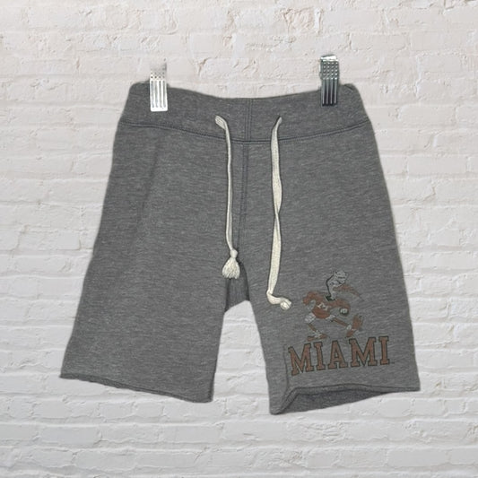 Grey shorts with white drawstrings featuring a Miami graphic logo, hanging against a brick wall.