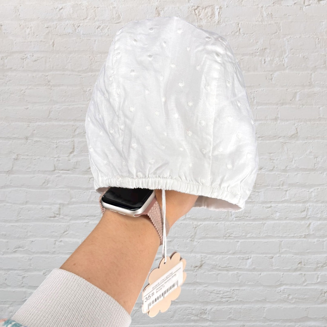 White vintage-style baby bonnet with delicate embroidery, held in front of a brick wall background.