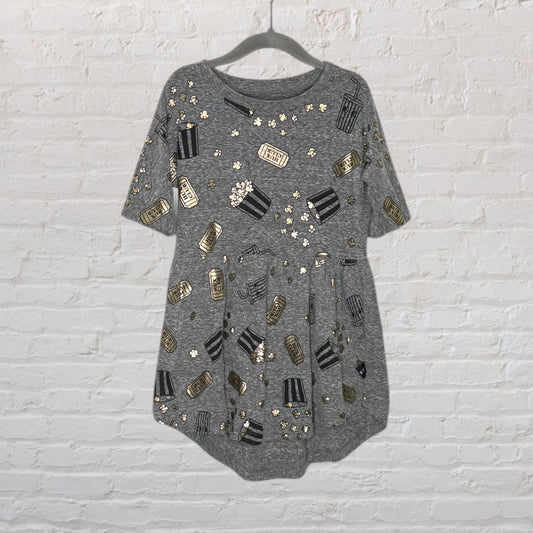 Grey tunic with black and gold popcorn and movie ticket print, short sleeves on a hanger against a white brick wall.