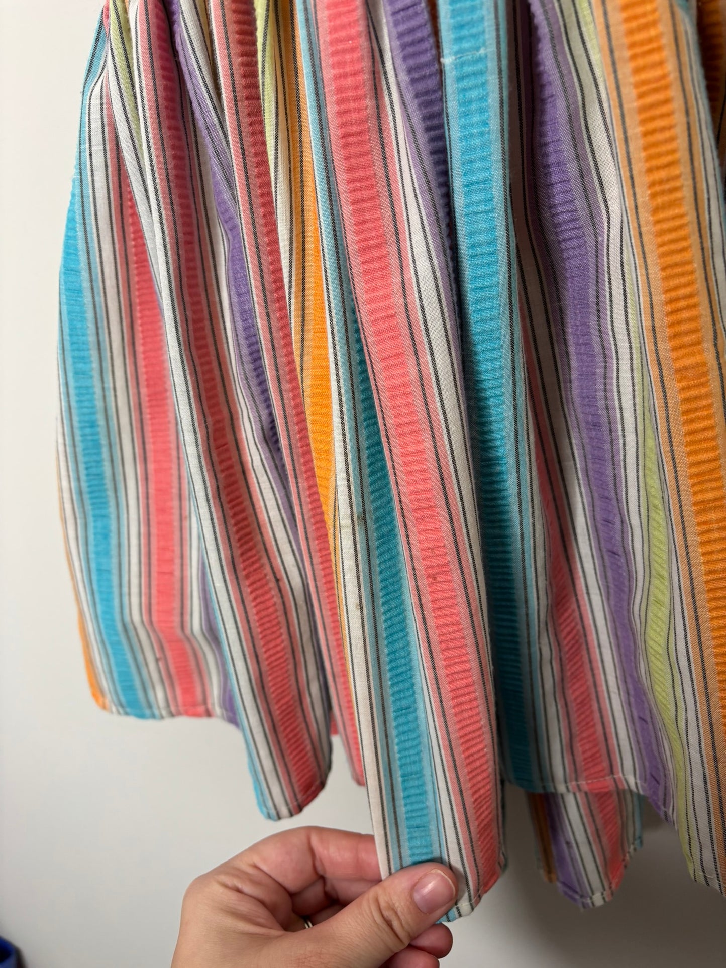 A vibrant rainbow-striped sundress with spaghetti straps, hanging on a black hanger against a white brick wall.