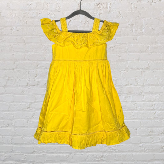 Yellow off-shoulder dress with ruffle details hanging on a hanger against a white brick wall.