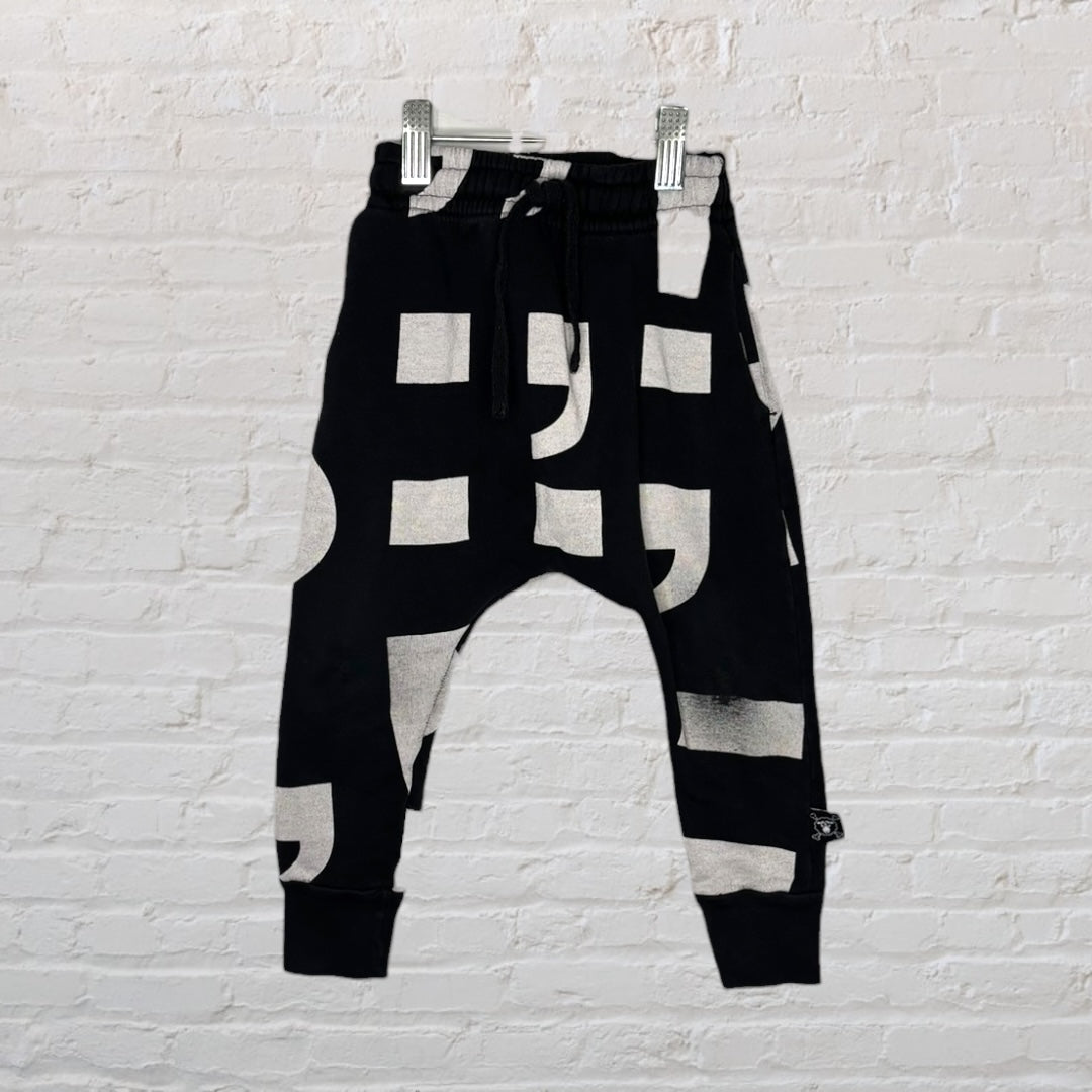 Black harem pants with bold white geometric patterns hanging on a hanger against a white brick wall.