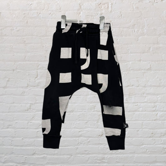 Black harem pants with bold white geometric patterns hanging on a hanger against a white brick wall.
