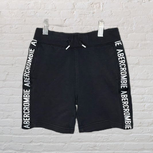 Black shorts with Abercrombie branding on the side strips displayed on a white brick wall.