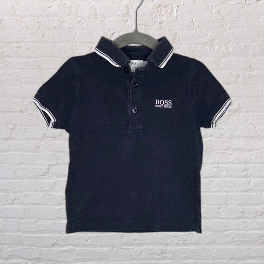 Navy blue Hugo Boss polo shirt with white logo and striped collar, displayed on a hanger.