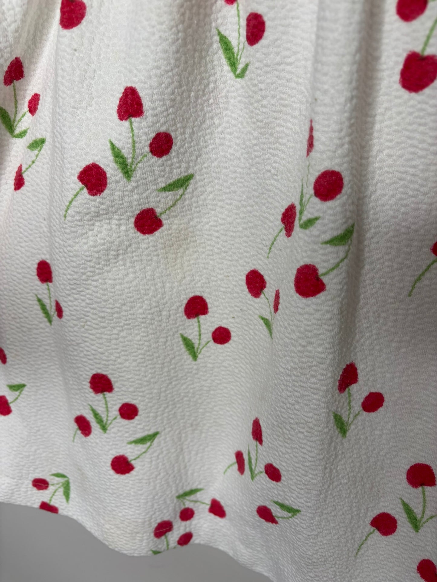 White dress with red cherry print and ruffled sleeves hanging on a black hanger against a textured wall.