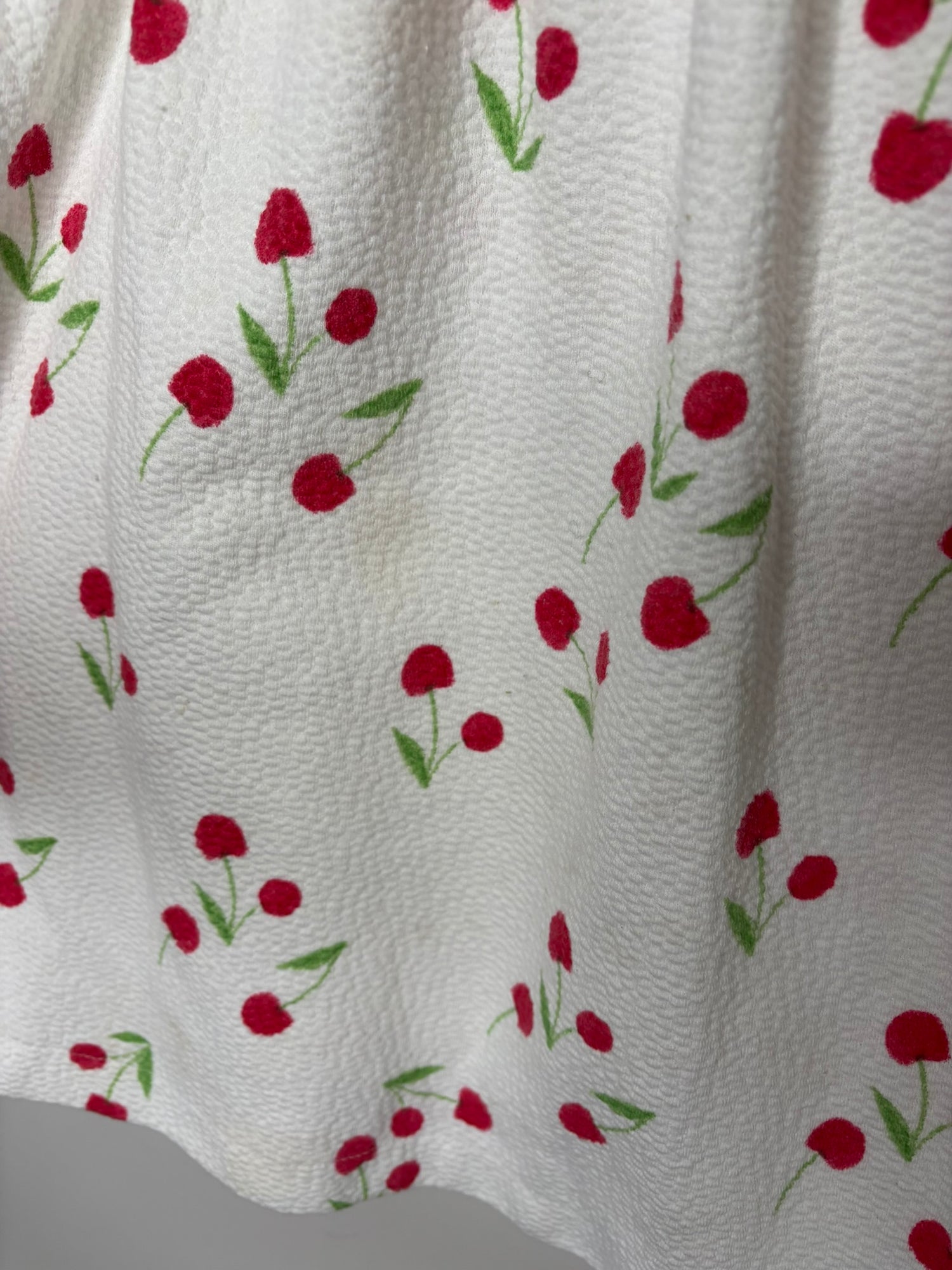 White dress with red cherry print and ruffled sleeves hanging on a black hanger against a textured wall.