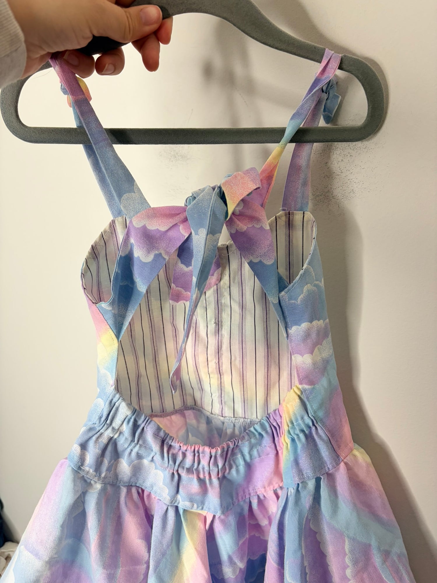 Children's dress with pastel rainbow and cloud print, featuring a ruffled hem and tied straps.