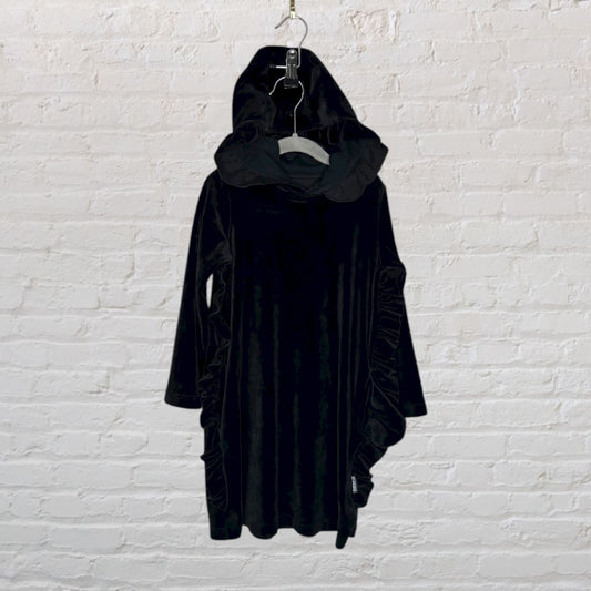 Black velvet dress with a hood and ruffled sides hanging against a white brick wall.