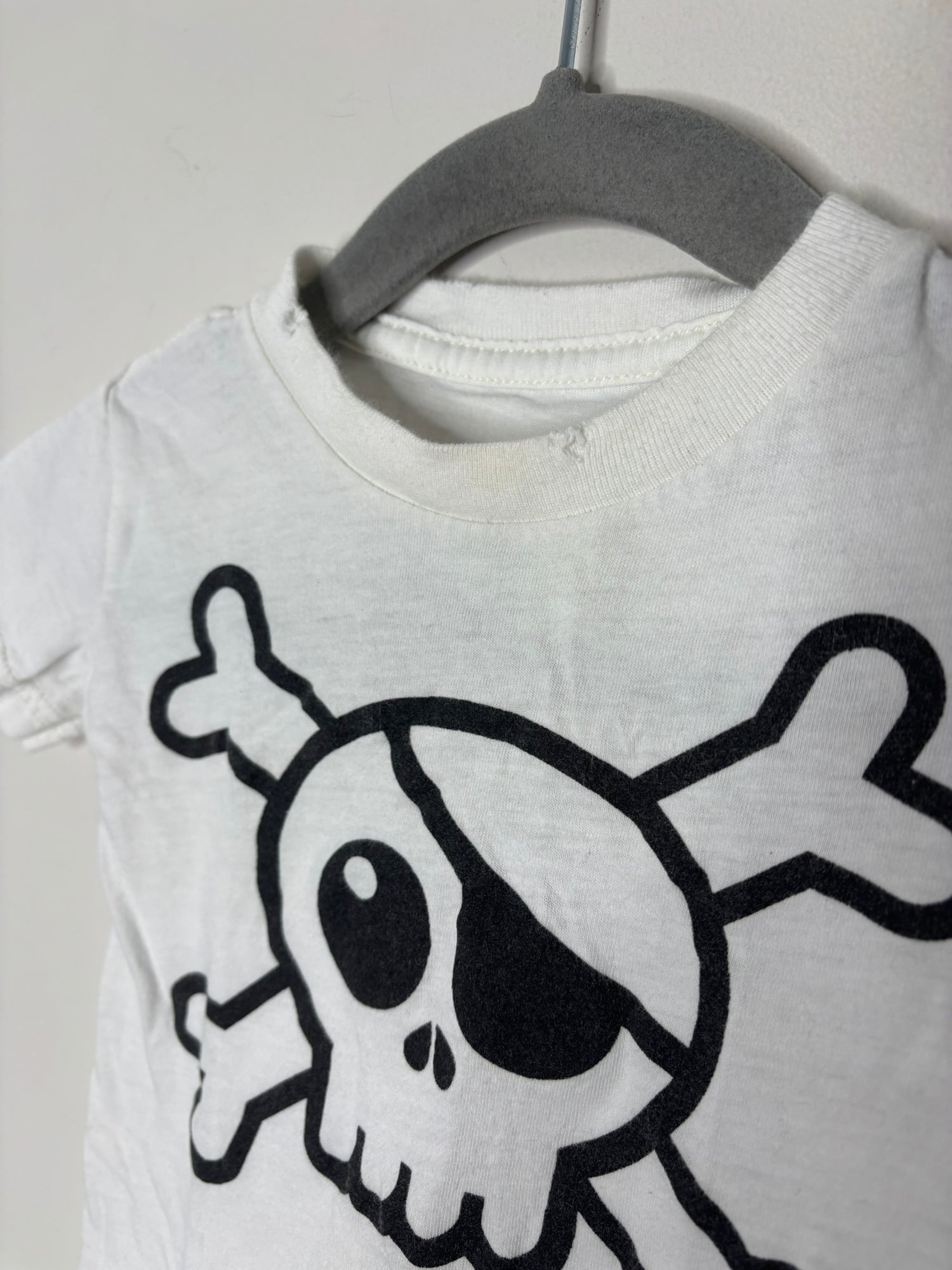 White T-shirt with a black skull and crossbones graphic, hanging on a hanger against a brick wall.