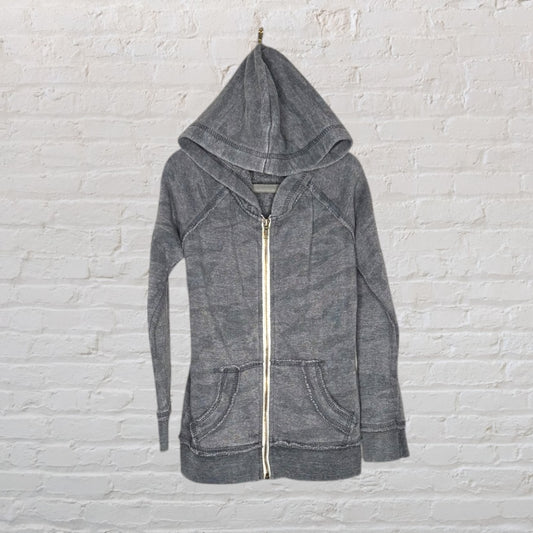 Gray washed zip-up hoodie with pockets and a hood, hanging against a white brick wall.